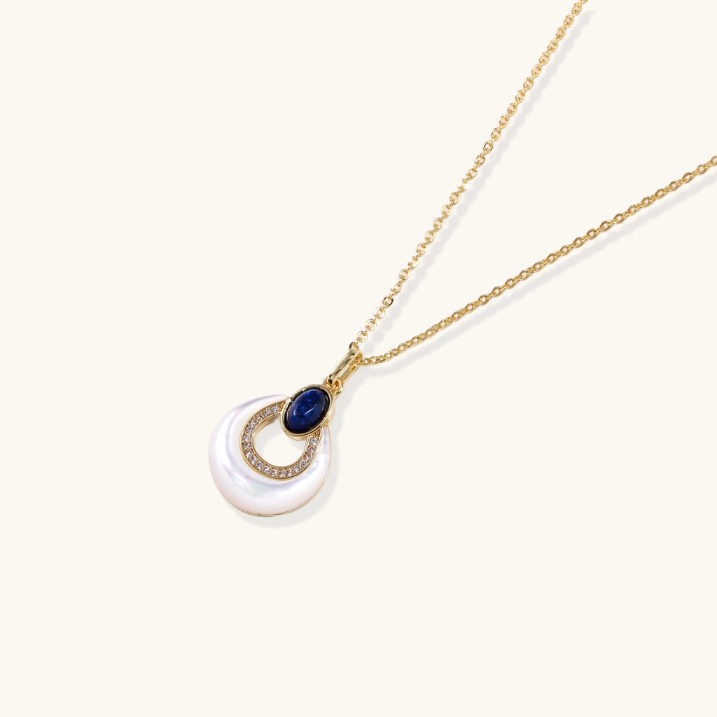 Gold plated chain necklace with a white MOP crescent pendant, blue lapis lazuli oval and sparkling stone accents on a cream background.