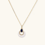 Gold plated chain necklace with a white MOP crescent pendant, blue lapis lazuli oval and sparkling stone accents on a cream background.