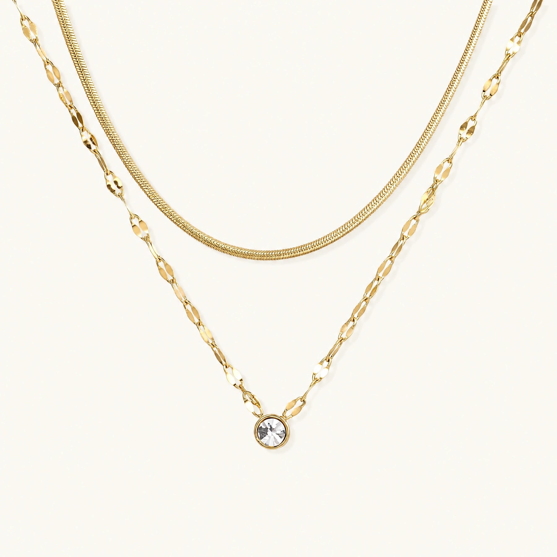 Layered gold necklace with herringbone and link chains, small bezel-set crystal pendant on cream background