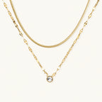 Layered gold necklace with herringbone and link chains, small bezel-set crystal pendant on cream background